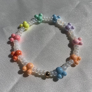 May include: A colorful beaded bracelet featuring pastel-colored Mickey Mouse-shaped beads. The bracelet includes beads in shades of pink, orange, yellow, green, blue, and purple, strung on a clear elastic cord. A small gold bead accents the design.