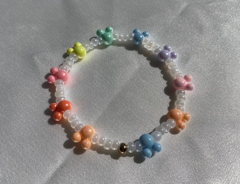 May include: A colorful beaded bracelet featuring pastel-colored Mickey Mouse-shaped beads. The bracelet includes beads in shades of pink, orange, yellow, green, blue, and purple, strung on a clear elastic cord. A small gold bead accents the design.