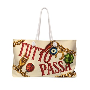 May include: A cream-colored tote bag with white rope handles featuring the red text TUTTO PASSA. The design includes gold chains, a red horn, a blue evil eye charm, a gold horseshoe, a green four-leaf clover, a ladybug, and a gold hand gesture.