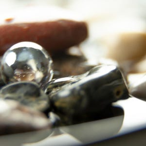 May include: Close-up of a collection of smooth, polished stones and a reflective silver sphere. The stones vary in color, including shades of brown, gray, and cream. The sphere reflects the surrounding objects.