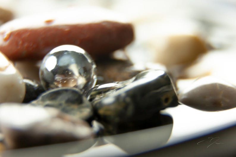 May include: Close-up of a collection of smooth, polished stones and a reflective silver sphere. The stones vary in color, including shades of brown, gray, and cream. The sphere reflects the surrounding objects.
