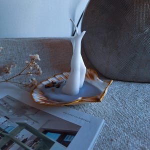 Op de afbeelding: Een witte kaars in de vorm van benen, geplaatst in een schelpvormige houder met gouden rand. De houder bevat een lichtblauwe vloeistof en kleine decoratieve stenen. Een tijdschrift met de tekst "OSLOBUKTA/BJØRVIKA" staat op de voorgrond.