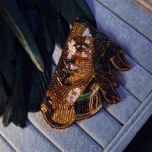 May include: A detailed brooch with a portrait design, crafted from gold, black, and white beads. The beaded artwork includes a green accent. The brooch is set against a grey background, showcasing the intricate beadwork.