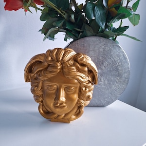 May include: A gold-colored, detailed sculpture of a woman's face with curly hair. The sculpture is on a white surface.