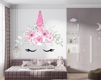 May include: A pink unicorn head wall decal with a floral crown and closed eyes. The decal is on a white wall in a bedroom with a pink couch and a white teddy bear.