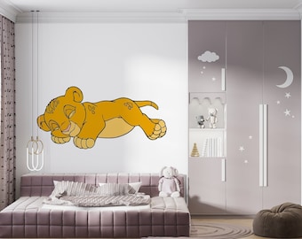 May include: A white wall with a cartoon illustration of a sleeping lion cub. The lion cub is orange and has closed eyes. The wall is decorated with a white cloud and stars. There is a white cupboard with a white moon and stars on the door. A brown beanbag chair is in the foreground.