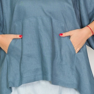 May include: A blue-grey linen top with a relaxed fit and a front pocket. The top has wide sleeves and a slightly asymmetrical hemline. The person's hands are in the pockets, with red nail polish.