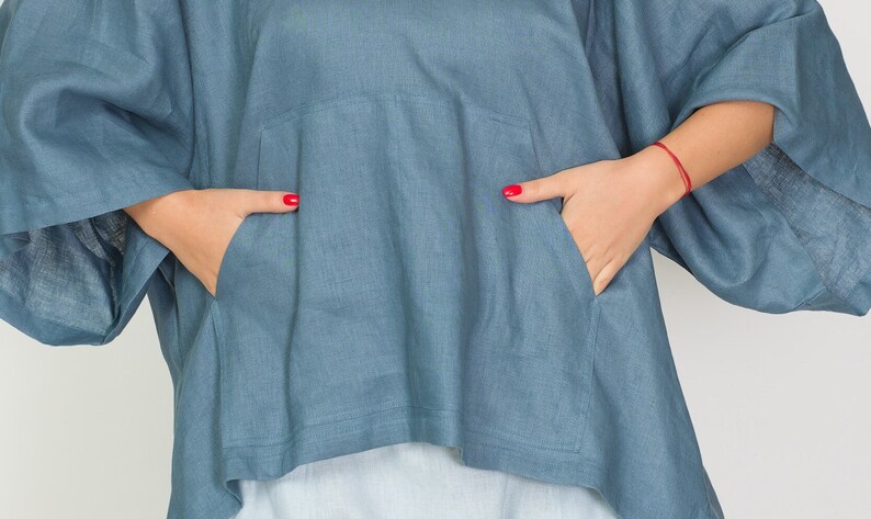 May include: A blue-grey linen top with a relaxed fit and a front pocket. The top has wide sleeves and a slightly asymmetrical hemline. The person's hands are in the pockets, with red nail polish.
