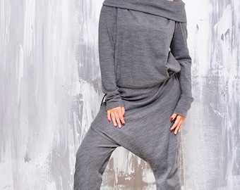 May include: A charcoal gray jumpsuit featuring a wide, folded off-the-shoulder neckline, long sleeves, and a relaxed, drop-crotch silhouette. The garment is crafted from a soft, heathered knit fabric and is paired with black lace-up boots.