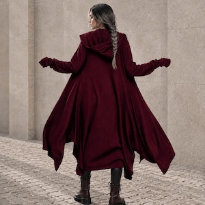 Könnte beinhalten: Eine langärmelige, tiefburgunderrote Kapuzenstrickjacke mit asymmetrischem, fließendem Saum und verlängerten Ärmelbündchen mit Daumenlöchern. Das Kleidungsstück wird über einer dunklen Hose und dunkelbraunen Schnürstiefeln getragen.