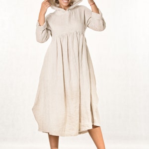 Op de afbeelding: Een beige midi-jurk met capuchon en lange mouwen, voorzien van een geplooide taille die een volle, zwierige rok creëert. De jurk is gemaakt van een textuurrijke stof en wordt gecombineerd met bijpassende beige veterschoenen met dikke zolen.