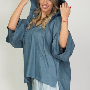 May include: A blue hooded poncho-style top with a V-neck and side slits. The top has wide sleeves and a front pocket. The model is wearing light blue pants. The outfit is made of a soft, flowing fabric.