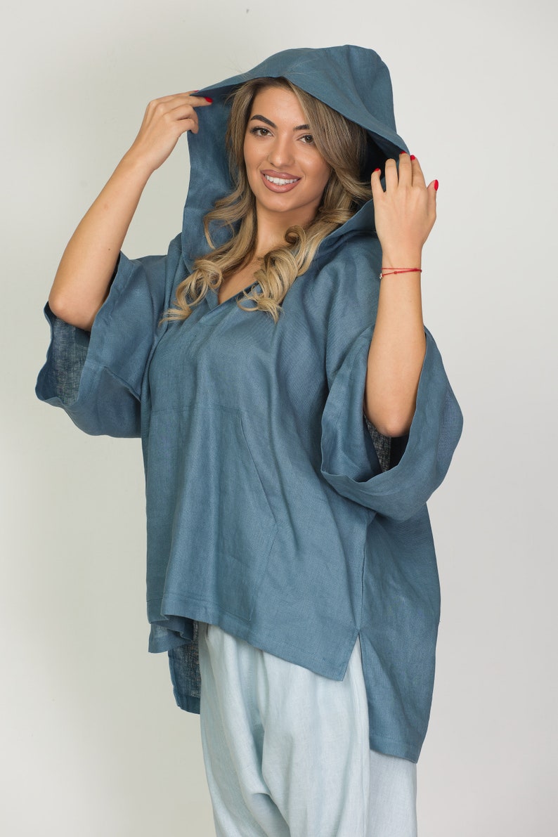 May include: A blue hooded poncho-style top with a V-neck and side slits. The top has wide sleeves and a front pocket. The model is wearing light blue pants. The outfit is made of a soft, flowing fabric.