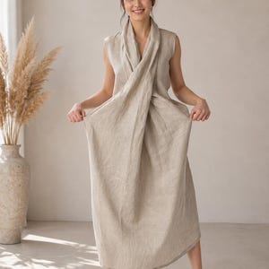 Op de afbeelding: Een mouwloze, enkellange beige linnen jurk met een gedrapeerd voorpand in wikkelstijl dat een gelaagd effect creëert. Het kledingstuk heeft een hoge halslijn en een los, vloeiend silhouet dat tot aan de enkels reikt, gemaakt van textuurrijke stof.