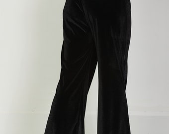 May include: Black velvet trousers featuring a high-waisted design with a wide-leg silhouette. The pants have a smooth, light-reflecting texture and are paired with black block-heeled boots and a dark green top tucked into the waistband.
