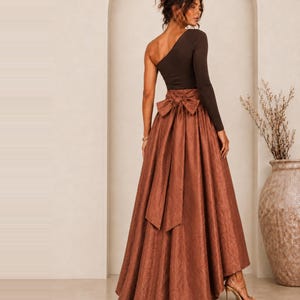 Peut inclure: Un haut à manches longues et une épaule dénudée marron foncé associé à une jupe longue taille haute de couleur cuivre. La jupe présente un tissu texturé, une large ceinture et un grand nœud décoratif noué au milieu du dos avec de longs pans.