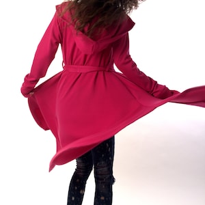 May include: A vibrant magenta hooded coat with long sleeves and a waist tie, worn over dark blue distressed denim jeans and white and black athletic sneakers. The coat features a flared hemline that extends outward during movement.