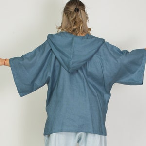 May include: A blue hooded kimono-style top with wide sleeves. The top is made of a lightweight fabric and has a relaxed fit. The hood is attached to the back of the top. The sleeves are wide and extend to the elbows. The top is a solid blue color.