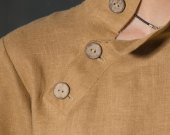 May include: Close-up of a tan-colored linen shirt with a mandarin collar. The shirt features three large, round, wooden buttons. The fabric has a textured appearance. The collar is buttoned at the neck.