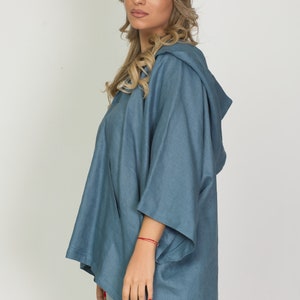 May include: A denim-style hooded top in a muted blue hue. The top features a relaxed fit with wide sleeves and a hood. The garment has a split hem and is made of a soft, flowing fabric, suitable for casual wear.
