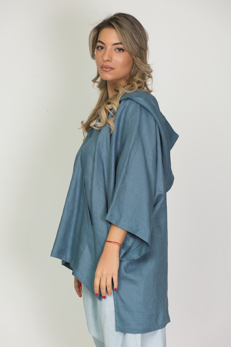 May include: A denim-style hooded top in a muted blue hue. The top features a relaxed fit with wide sleeves and a hood. The garment has a split hem and is made of a soft, flowing fabric, suitable for casual wear.