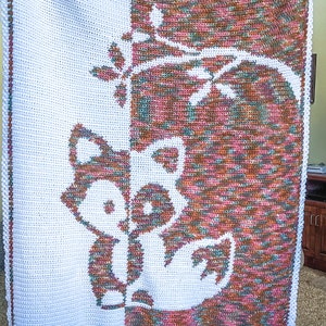 Könnte beinhalten: Eine weiße Häkeldecke mit einem braunen und rosafarbenen Waschbär-Design. Der Waschbär ist weiß umrandet und der Hintergrund ist ein braunes und rosafarbenes Ombre.