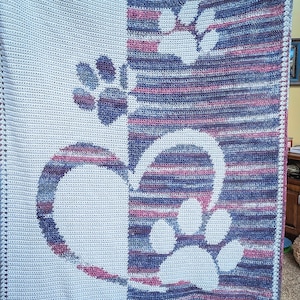 May include: A crocheted blanket with a white background and a purple and blue striped pattern. The blanket features a design of paw prints in white.