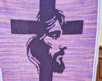 May include: A crocheted blanket in shades of purple, lavender, and white features a large, dark purple cross with a silhouette of a person praying. The blanket has a textured appearance and a white border.