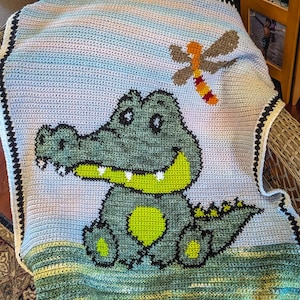 May include: Crocheted baby blanket featuring a friendly alligator design in shades of green and yellow. The blanket has a striped background in pastel colors and a dragonfly applique. The edges are finished with a black and white border.