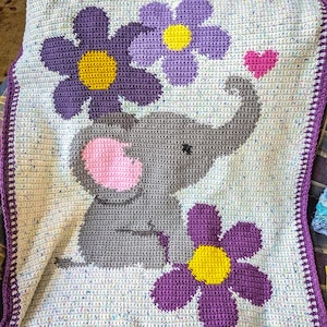 May include: A crocheted baby blanket with a gray elephant and purple flowers. The blanket has a white border with a crocheted edge.