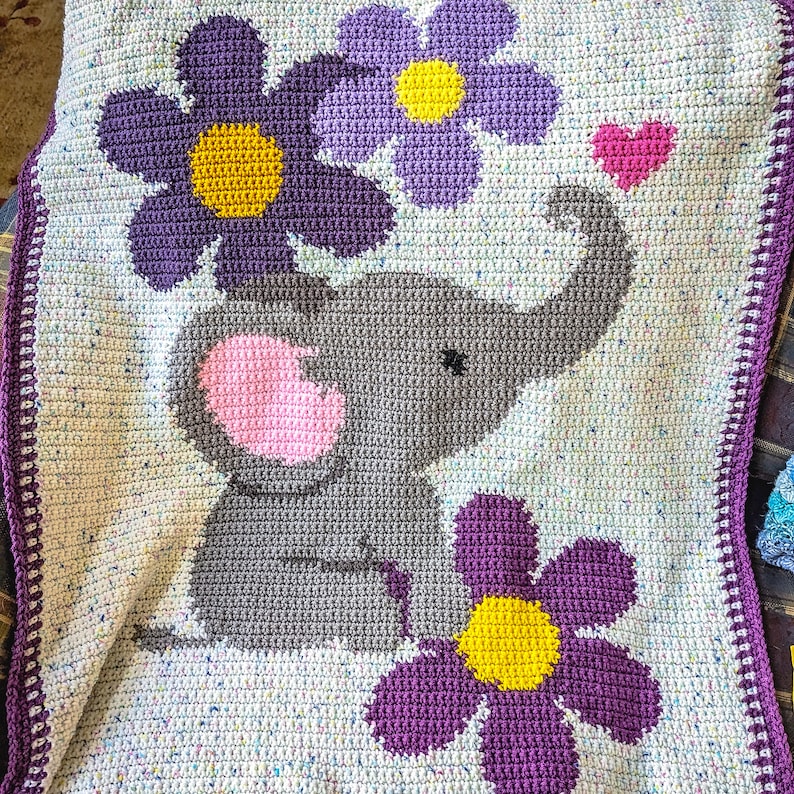May include: A crocheted baby blanket with a gray elephant and purple flowers. The blanket has a white border with a crocheted edge.
