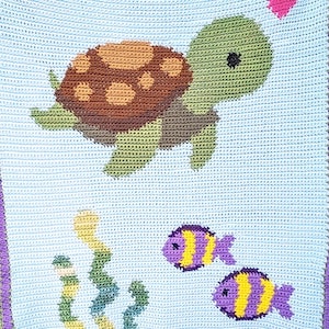 Puede incluir: Manta de ganchillo con una tortuga marina con caparazón marrón y cabeza y aletas verdes, nadando sobre dos peces morados y amarillos. El fondo es azul claro, con algas verdes y un corazón rosa.
