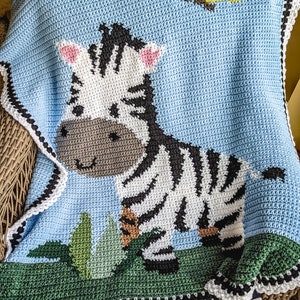 Pode incluir: Um cobertor de bebé em crochê com um zebra com listras pretas e brancas, rodeado de folhas verdes sobre um fundo azul claro.