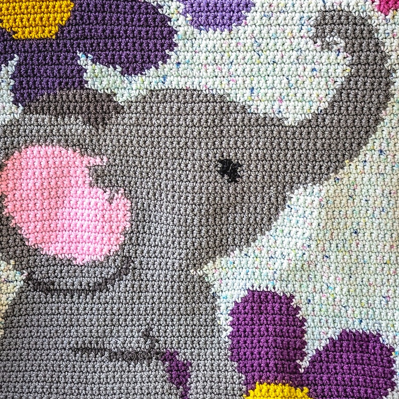 May include: A crocheted baby blanket with a gray elephant design, pink nose, and black eye. The blanket is made with a variety of colors, including purple, yellow, and white.