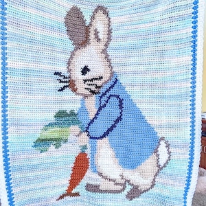 May include: A crocheted blanket featuring Peter Rabbit holding a carrot. The blanket has a blue and white striped background with a blue border. Peter Rabbit is depicted in white, brown, and blue, with a carrot in his hand.