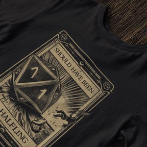 May include: A black t-shirt with a graphic of a dice with the number 7 on it. The graphic is surrounded by a border with the text "Should Have Been A Halfling".