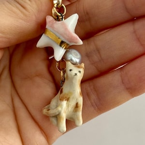 May include: A gold-toned metal lobster clasp charm featuring a white and pink ceramic star with a gold band, a small grey pearl bead, and a white and brown spotted ceramic cat figurine hanging from a series of gold-toned metal jump rings.