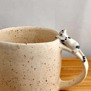 May include: A cream-colored ceramic mug with a speckled finish and a handle shaped like a small white cat with black spots. The cat is positioned at the top of the handle, appearing to rest its front paws on the rim of the mug while looking down.