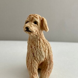 May include: A small ceramic sculpture of a dog sitting with its head tilted up. The dog is light brown and has a textured surface.