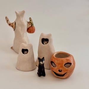 May include: A collection of ceramic figurines including a tall white ghost figure holding an orange pumpkin, two smaller white ghost figures with black cats peeking through openings, a small black cat figurine, and an orange jack-o'-lantern bowl.