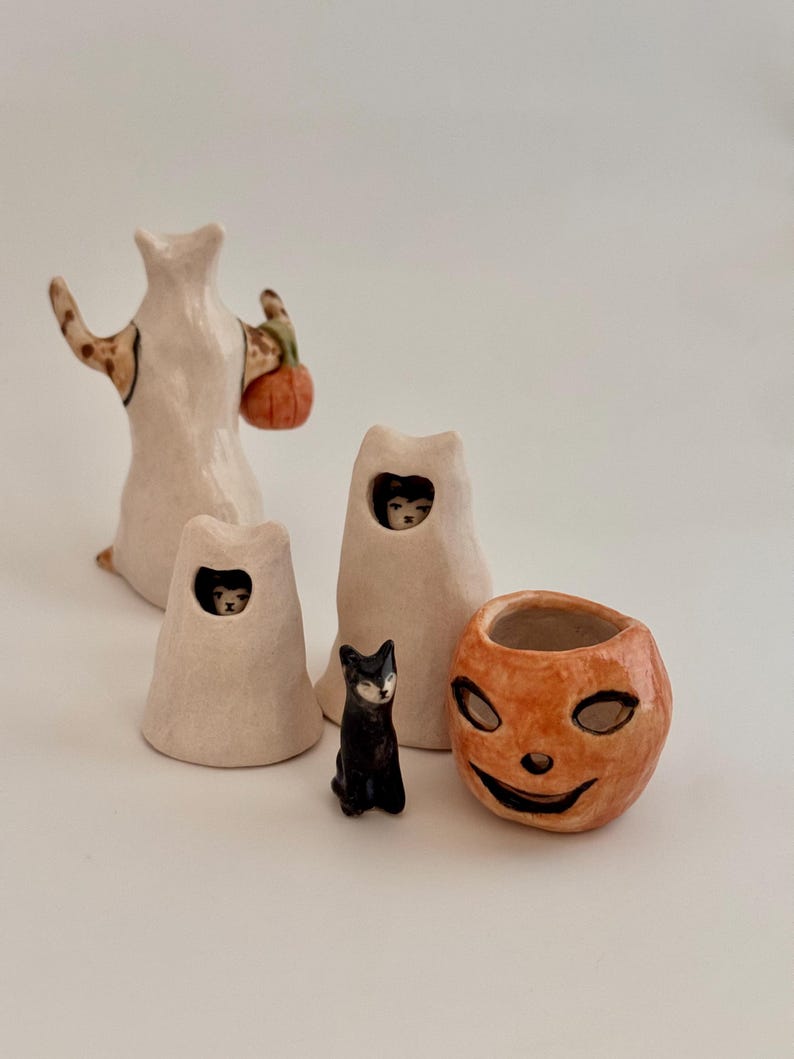 May include: A collection of ceramic figurines including a tall white ghost figure holding an orange pumpkin, two smaller white ghost figures with black cats peeking through openings, a small black cat figurine, and an orange jack-o'-lantern bowl.