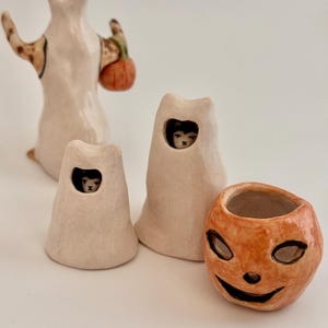 May include: A collection of four ceramic figurines including a tall ghost holding an orange pumpkin, two smaller ghost figures with visible cat faces peering through cutouts, and a separate orange jack-o'-lantern bowl with a carved face.