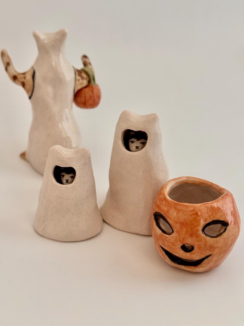 May include: A collection of four ceramic figurines including a tall ghost holding an orange pumpkin, two smaller ghost figures with visible cat faces peering through cutouts, and a separate orange jack-o'-lantern bowl with a carved face.