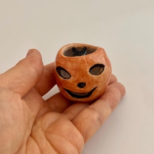 May include: A small, round, orange ceramic jack-o'-lantern vessel with a glossy finish. The pumpkin features hand-painted black eyes, a small nose, and a wide, smiling mouth. A tiny, sculpted black cat figure is visible inside the hollow opening.
