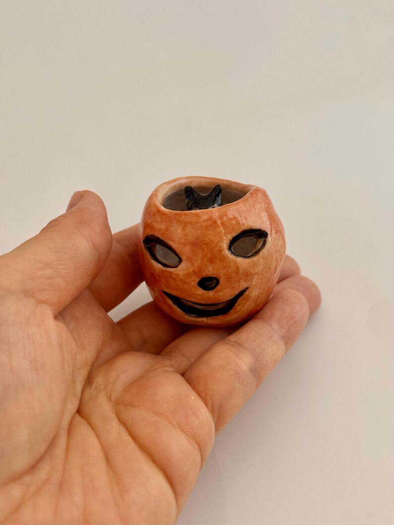 May include: A small, round, orange ceramic jack-o'-lantern vessel with a glossy finish. The pumpkin features hand-painted black eyes, a small nose, and a wide, smiling mouth. A tiny, sculpted black cat figure is visible inside the hollow opening.