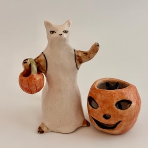 May include: A ceramic figurine of a white cat with brown markings wearing a white garment and holding a small orange pumpkin bucket, positioned next to a separate orange ceramic jack-o'-lantern container with a black interior.