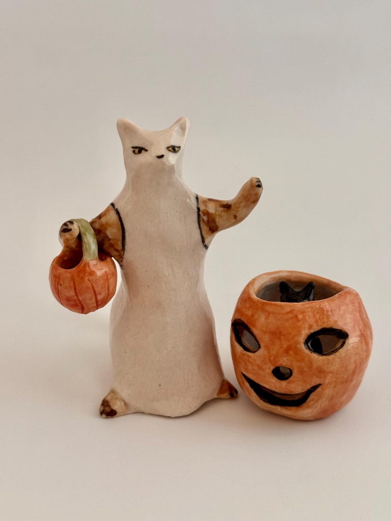 May include: A ceramic figurine of a white cat with brown markings wearing a white garment and holding a small orange pumpkin bucket, positioned next to a separate orange ceramic jack-o'-lantern container with a black interior.