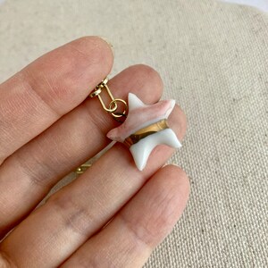 Pu&ograve; includere: Un orecchino a forma di stella con un gancio in metallo color oro. La stella &egrave; bianca con accenti rosa e una fascia dorata al centro. L'orecchino &egrave; tenuto in mano da una persona.