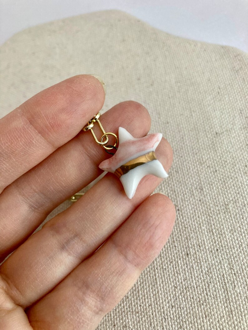 Pu&ograve; includere: Un orecchino a forma di stella con un gancio in metallo color oro. La stella &egrave; bianca con accenti rosa e una fascia dorata al centro. L'orecchino &egrave; tenuto in mano da una persona.