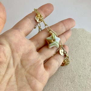 Pu&ograve; includere: Bracciale a catena dorata con un ciondolo a forma di stella a strisce verdi e bianche e dettagli dorati. Il bracciale include anche piccoli ciondoli rotondi dorati. Il gioiello &egrave; presentato su uno sfondo neutro.